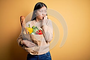 Young asian woman holding paper bag of fresh healthy groceries over yellow isolated background looking stressed and nervous with