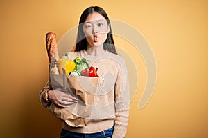 Young asian woman holding paper bag of fresh healthy groceries over yellow isolated background making fish face with lips, crazy