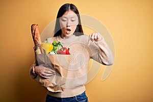 Young asian woman holding paper bag of fresh healthy groceries over yellow isolated background Pointing down with fingers showing