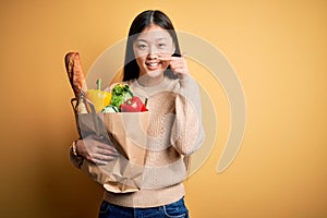 Young asian woman holding paper bag of fresh healthy groceries over yellow isolated background Pointing with hand finger to face