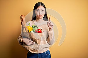 Young asian woman holding paper bag of fresh healthy groceries over yellow isolated background Pointing up looking sad and upset,