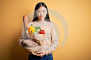 Young asian woman holding paper bag of fresh healthy groceries over yellow isolated background Relaxed with serious expression on