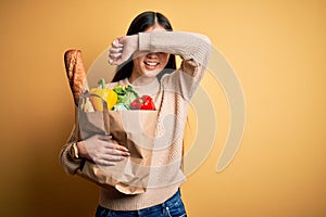 Young asian woman holding paper bag of fresh healthy groceries over yellow isolated background Smiling cheerful playing peek a boo