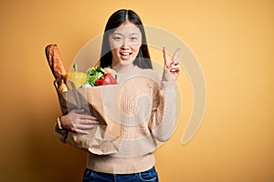 Young asian woman holding paper bag of fresh healthy groceries over yellow isolated background smiling with happy face winking at