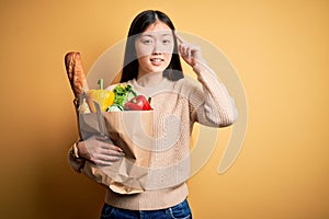 Young asian woman holding paper bag of fresh healthy groceries over yellow isolated background Smiling pointing to head with one