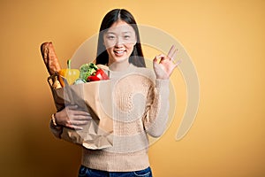 Young asian woman holding paper bag of fresh healthy groceries over yellow isolated background smiling positive doing ok sign with