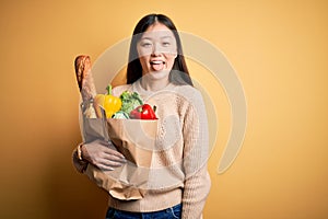 Young asian woman holding paper bag of fresh healthy groceries over yellow isolated background sticking tongue out happy with