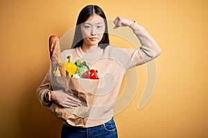 Young asian woman holding paper bag of fresh healthy groceries over yellow isolated background Strong person showing arm muscle,