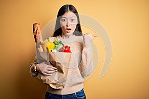 Young asian woman holding paper bag of fresh healthy groceries over yellow isolated background Surprised pointing with hand finger