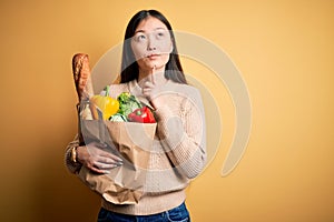 Young asian woman holding paper bag of fresh healthy groceries over yellow isolated background Thinking concentrated about doubt