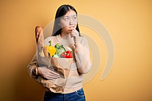 Young asian woman holding paper bag of fresh healthy groceries over yellow isolated background Thinking worried about a question,