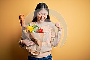 Young asian woman holding paper bag of fresh healthy groceries over yellow isolated background very happy and excited doing winner