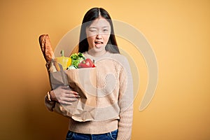 Young asian woman holding paper bag of fresh healthy groceries over yellow isolated background winking looking at the camera with