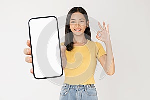 Young Asian woman holding smartphone with blank screen for advertising and showing ok-sign on white background