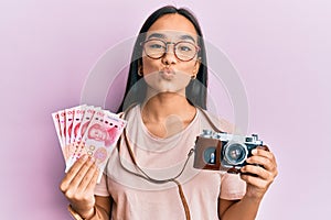 Young asian woman holding vintage camera and 100 chinese yuan looking at the camera blowing a kiss being lovely and sexy