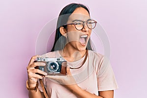 Young asian woman holding vintage camera angry and mad screaming frustrated and furious, shouting with anger