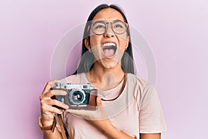 Young asian woman holding vintage camera angry and mad screaming frustrated and furious, shouting with anger looking up
