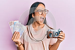 Young asian woman holding vintage camera and swedish krona angry and mad screaming frustrated and furious, shouting with anger