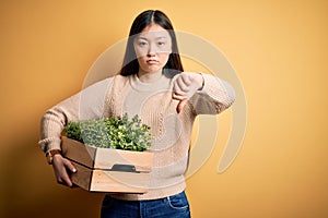 Young asian woman holding wood box of fresh garden natural plants over yellow background with angry face, negative sign showing
