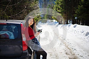 Young asian woman holds mobile phone near car on a winter road. Cold weather, snow-covered trees in the winter forest.