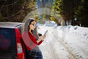 Young asian woman holds mobile phone near car on a winter road. Cold weather, snow-covered trees in the winter forest.