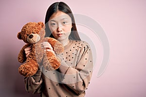 Young asian woman hugging teddy bear stuffed animal over pink  background with a confident expression on smart face