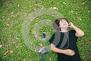 Young Asian woman laying on the green grass listening to music in the park with a chill emotion.