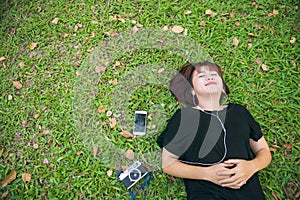 Young Asian woman laying on the green grass listening to music in the park with a chill emotion.