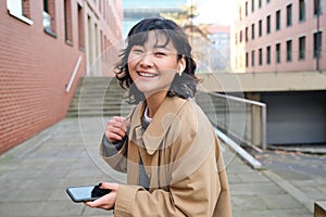 Young asian woman listens music in wireless earphones, uses smartphone on street, walks around city on sunny day