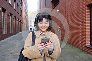 Young asian woman listens music in wireless earphones, uses smartphone on street, walks around city on sunny day