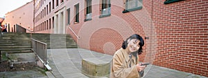 Young asian woman listens music in wireless earphones, uses smartphone on street, walks around city on sunny day