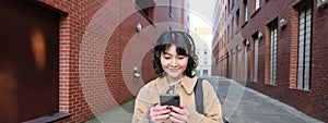 Young asian woman listens music in wireless earphones, uses smartphone on street, walks around city on sunny day