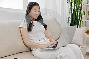 Young Asian woman looking at laptop screen on her lap sitting on couch at home lifestyle work as a freelancer