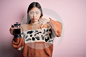 Young asian woman making a glass of coffe using french press coffee maker over pink background with angry face, negative sign