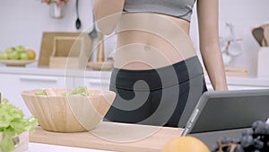 Young Asian woman making salad healthy food while using tablet for looking recipe and drinking orange juice in the kitchen.