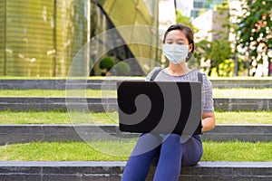 Young Asian woman with mask for protection from corona virus outbreak using laptop while sitting outdoors