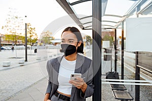Young asian woman in medical face mask using cellphone while standing at bus station