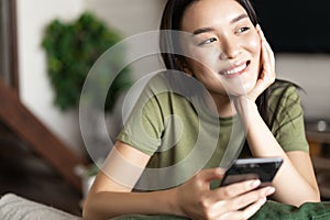 Young asian woman placing an order, waiting for delivery on mobile phone app, sitting at home and looking outside window