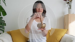 Young Asian woman playing Tibetan singing bowl sitting in meditation couch at home. 4k resolution.