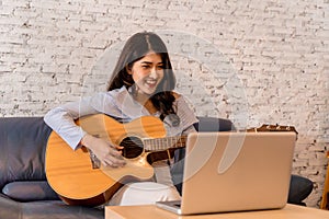 Young Asian woman practicing and learning how to play guitar on laptop computer monitor. Female guitarist watching photo