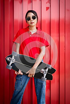 Young asian woman in red T-shirt with collar holding the surfskate board in hip position in front of red galvanized steel sheet