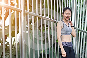 Young asian woman relaxing after jogging exercise stand against steel in park to freshen her body and enjoy warm light in morning.