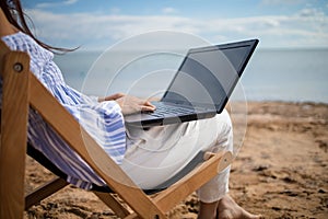 Young asian woman is resting on a lounger with a laptop in her hands on a sandy sunny beach near the ocean. Freelance and remote w