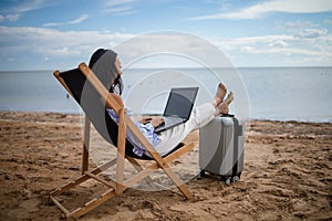 Young asian woman is resting on a lounger with a laptop in her hands on a sandy sunny beach near the ocean. Freelance and remote w