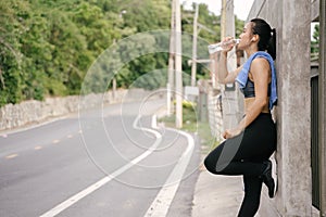 Young Asian woman runner resting after workout running and drinking water in street road.