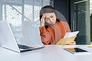 A young Asian woman, secretary manager, accountant is worriedly holding a letter, document, account. She sits confused