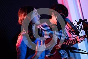 Young asian woman singing while man in background playing acoustic guitar.