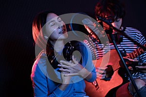 Young asian woman singing while man in background playing acoustic guitar.