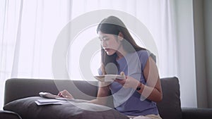 Young asian woman sit on sofa using a calculator to calculate income and expenses or managing monthly budget