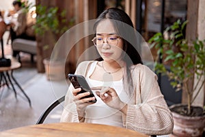 A young Asian woman sits at an outdoor table of a cafe and uses her phone to read something online